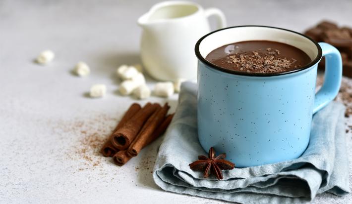 Imagem de uma caneca azul com chocolate quente. Na foto, pedaços de canela em pau, anis estrelado e uma mini jarra branca.